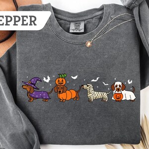 May include: Dark gray sweatshirt with a Halloween-themed dachshund design. The graphic features four dachshunds dressed as a witch, a pumpkin, a mummy, and a ghost, with bats in the background. The word "PEPPER" is in a white box in the upper left corner.