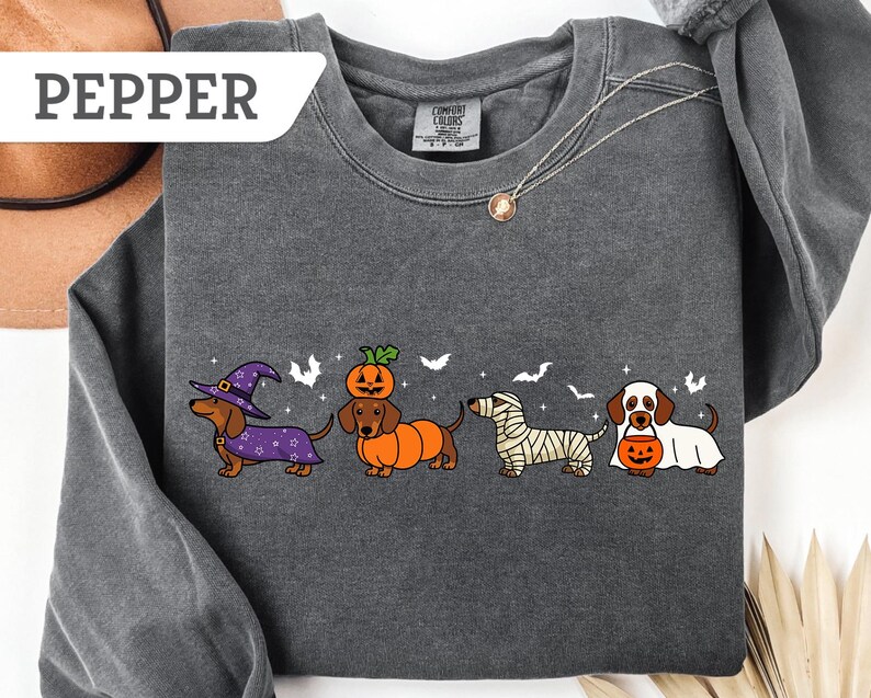 May include: Dark gray sweatshirt with a Halloween-themed dachshund design. The graphic features four dachshunds dressed as a witch, a pumpkin, a mummy, and a ghost, with bats in the background. The word "PEPPER" is in a white box in the upper left corner.