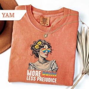 May include: Coral-colored t-shirt featuring a graphic of a woman with rainbow heart-shaped sunglasses and yellow flowers in her hair. The text "MORE PRIDE LESS PREJUDICE" is printed below the image. A gold necklace is draped across the shirt.