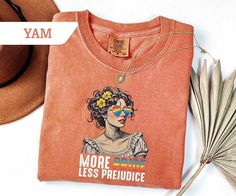 May include: Coral-colored t-shirt featuring a graphic of a woman with rainbow heart-shaped sunglasses and yellow flowers in her hair. The text "MORE PRIDE LESS PREJUDICE" is printed below the image. A gold necklace is draped across the shirt.
