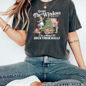 May include: Dark gray t-shirt featuring a Christmas-themed graphic. The design includes the text "From The Windows to The Walls, I'm About To DECK THESE HALLS" with illustrations of children decorating a Christmas tree and presents.