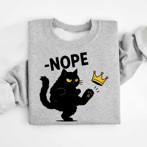 May include: A grey sweatshirt featuring a black cat kicking a gold crown. The word "-NOPE" is above the cat. The cat has yellow eyes and is kicking the crown with its paw.