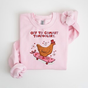 K&ouml;nnte beinhalten: Hellrosa Sweatshirt mit dem Text "OFF TO COMMIT TOMFOOLERY" in Rot. Das Design zeigt ein Cartoon-Huhn mit Zylinder auf einem rosa Skateboard mit Herzen und B&auml;ndern. Ein lustiges, skurriles Design.