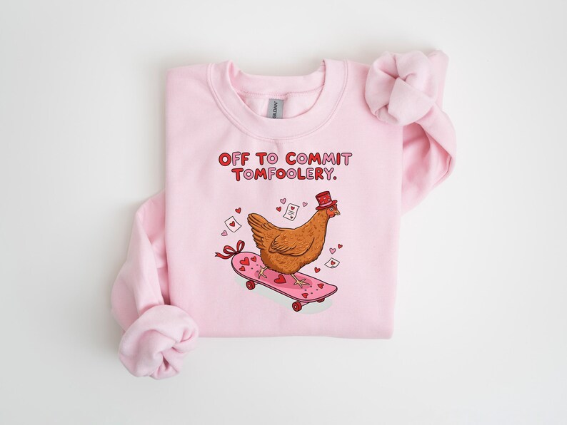 K&ouml;nnte beinhalten: Hellrosa Sweatshirt mit dem Text "OFF TO COMMIT TOMFOOLERY" in Rot. Das Design zeigt ein Cartoon-Huhn mit Zylinder auf einem rosa Skateboard mit Herzen und B&auml;ndern. Ein lustiges, skurriles Design.