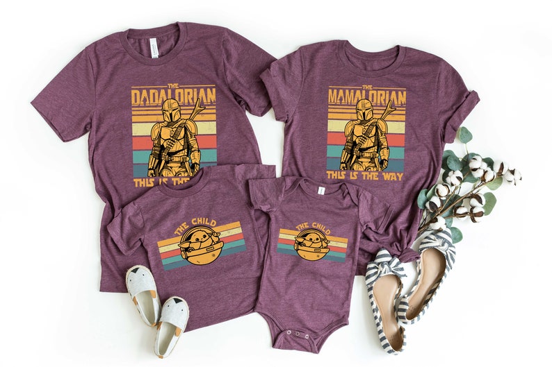 May include: A set of four maroon t-shirts with a retro-style graphic of a Mandalorian character. The shirts feature the text "The Dadalorian", "The Mamalorian", and "The Child".