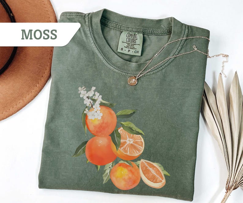 May include: Moss green t-shirt with a crew neck and short sleeves. The shirt features a watercolor illustration of oranges with green leaves and white flowers. A gold necklace with a pendant is draped on the shirt.