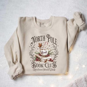 May include: A beige sweatshirt with a graphic design of a mug of hot cocoa with a gingerbread man, books, and the text "North Pole Book Club Hot Cocoa Served Daily".