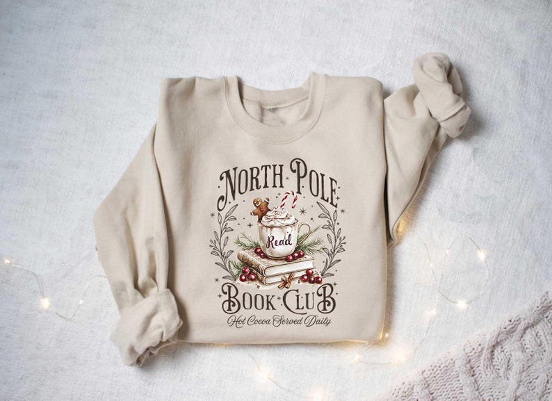 May include: A beige sweatshirt with a graphic design of a mug of hot cocoa with a gingerbread man, books, and the text "North Pole Book Club Hot Cocoa Served Daily".