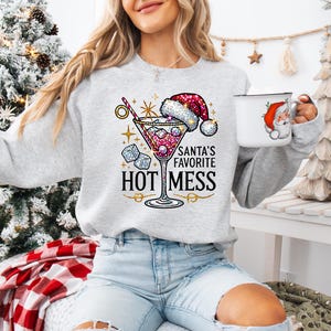 May include: A gray sweatshirt with a festive design featuring a martini glass with a red and white Santa hat, ice cubes, and gold stars. The text reads "Santa's Favorite Hot Mess".