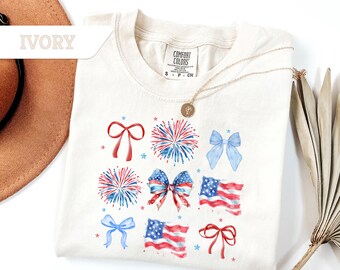 May include: A white t-shirt with a watercolor design of red, white, and blue bows, fireworks, and American flags. The shirt is labeled "Comfort Colors" and has a size tag that says "S - P - CH".