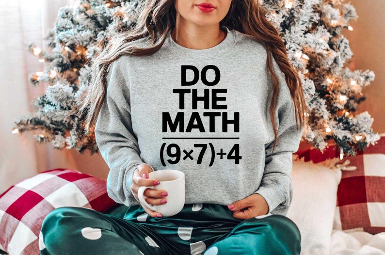 May include: A light gray sweatshirt with the text "DO THE MATH" above the equation (9x7)+4. The person is holding a white mug. The person is wearing green satin pants with white polka dots. A Christmas tree is in the background.