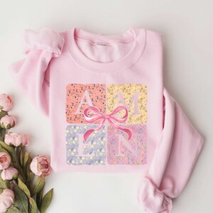 K&ouml;nnte beinhalten: Rosafarbener Pullover mit einem Blumendesign und dem Wort "AMEN" in der Mitte. Ein rosa Schleife befindet sich &uuml;ber dem Wort.