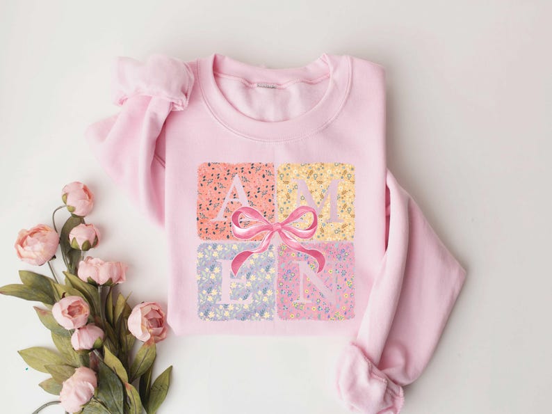K&ouml;nnte beinhalten: Rosafarbener Pullover mit einem Blumendesign und dem Wort "AMEN" in der Mitte. Ein rosa Schleife befindet sich &uuml;ber dem Wort.