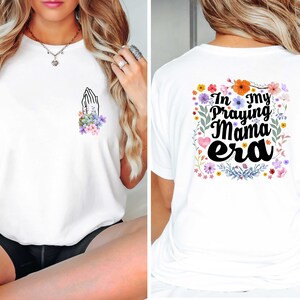 May include: White t-shirt with a floral design and the text "In my praying mama era" on the back. The front of the shirt has a small floral design with a praying hands graphic.