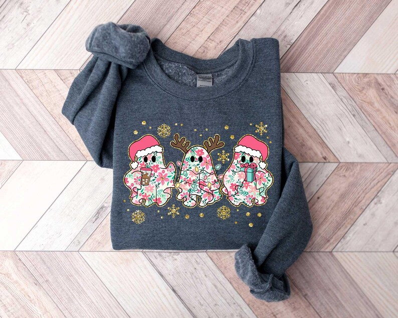 Puede incluir: Una sudadera gris oscuro con un dise&ntilde;o festivo que presenta tres fantasmas con gorros de Pap&aacute; Noel y sosteniendo regalos. Los fantasmas est&aacute;n decorados con patrones florales y rodeados de purpurina dorada.