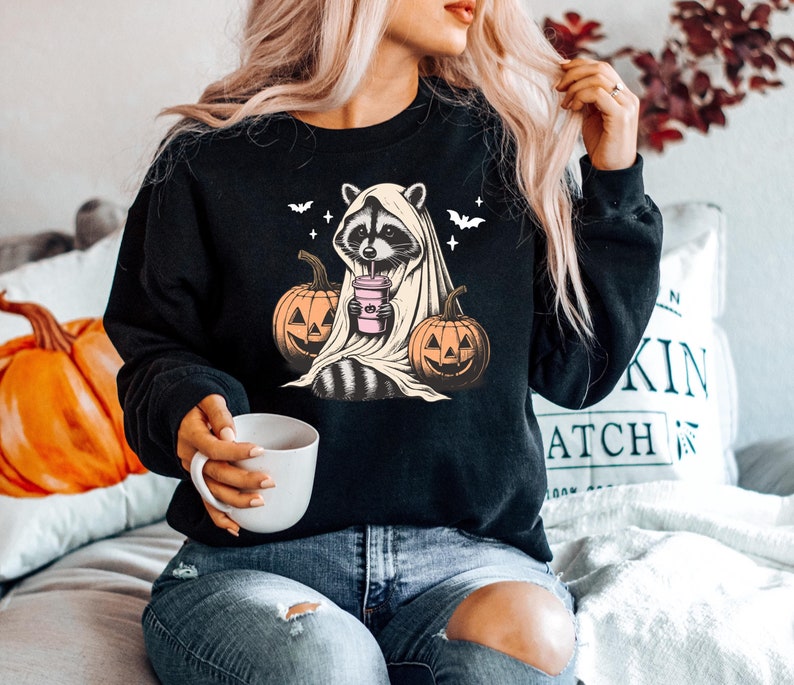 Puede incluir: Sudadera negra con un gr&aacute;fico de un mapache vestido de fantasma y sosteniendo una bebida rosa. El mapache est&aacute; rodeado de calabazas.