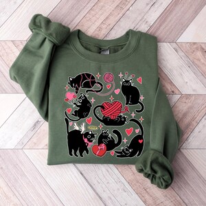 May include: A green sweatshirt with a black cat design. The cats are holding hearts, yarn, and a gift box. The design is surrounded by hearts and stars.