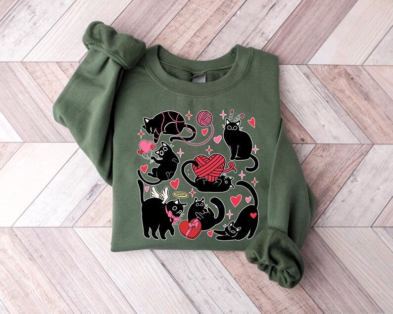 May include: A green sweatshirt with a black cat design. The cats are holding hearts, yarn, and a gift box. The design is surrounded by hearts and stars.