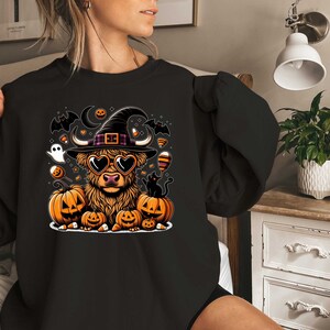 May include: A black sweatshirt with a Halloween-themed design featuring a Highland cow wearing a witch's hat and heart-shaped sunglasses. The cow is surrounded by pumpkins, bats, and candy corn.