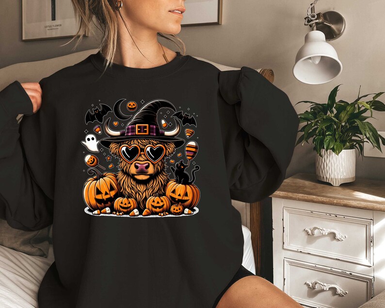 May include: A black sweatshirt with a Halloween-themed design featuring a Highland cow wearing a witch's hat and heart-shaped sunglasses. The cow is surrounded by pumpkins, bats, and candy corn.