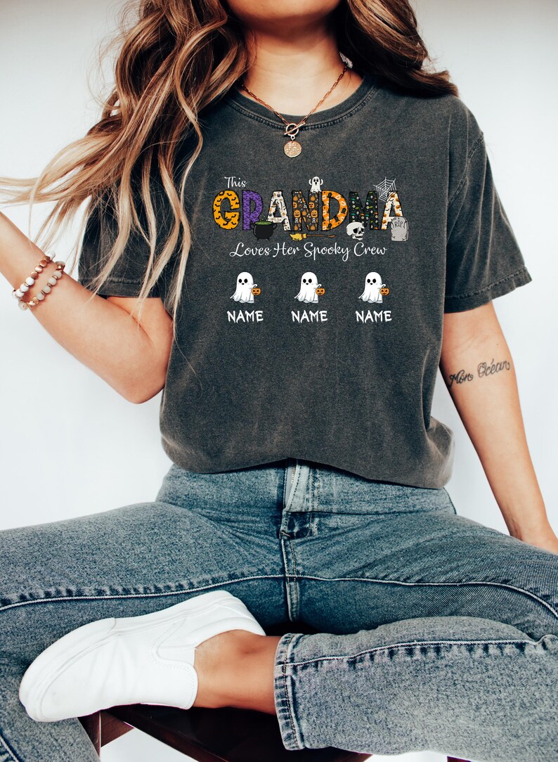 May include: Dark gray t-shirt with the text "This Grandma Loves Her Spooky Crew." The word "GRANDMA" is in orange, purple, and black Halloween-themed lettering. Below are three ghost illustrations with the word "NAME" under each.