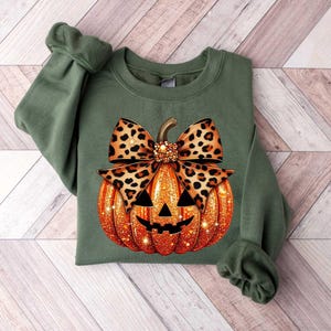 Peut inclure: Sweat-shirt vert avec un motif de citrouille sculpt&eacute;e. La citrouille est orange avec des paillettes et un n&oelig;ud en imprim&eacute; l&eacute;opard noir et marron sur le dessus.