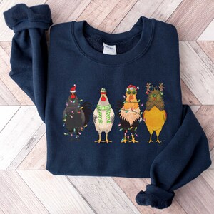 May include: A navy blue sweatshirt with a Christmas-themed design featuring four chickens wearing Santa hats and Christmas lights. The chickens are standing in a row and have different colored feathers.