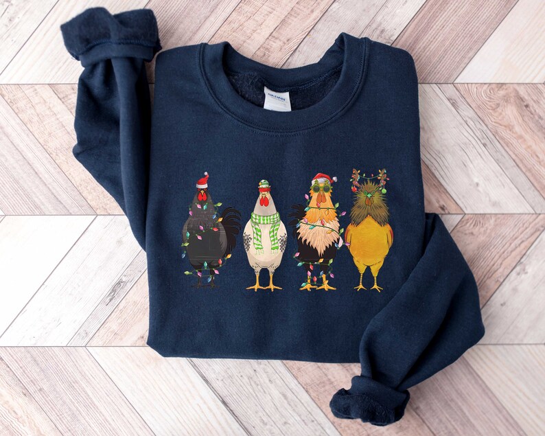 May include: A navy blue sweatshirt with a Christmas-themed design featuring four chickens wearing Santa hats and Christmas lights. The chickens are standing in a row and have different colored feathers.