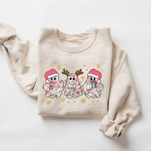 Puede incluir: Una sudadera beige con un dise&ntilde;o navide&ntilde;o con tres fantasmas que llevan gorros de Pap&aacute; Noel y sostienen regalos. Los fantasmas est&aacute;n decorados con patrones florales.