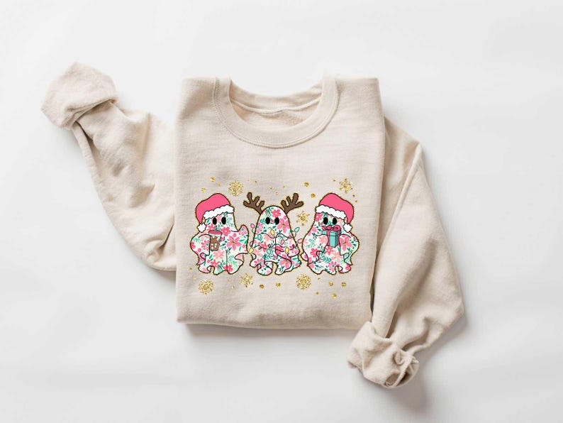 Puede incluir: Una sudadera beige con un dise&ntilde;o navide&ntilde;o con tres fantasmas que llevan gorros de Pap&aacute; Noel y sostienen regalos. Los fantasmas est&aacute;n decorados con patrones florales.