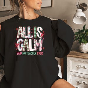 May include: Black sweatshirt with the text "ALL IS CALM" in white, green, and pink, with decorative elements. Below the text is the phrase "SAID NO TEACHER EVER" in white. The sweatshirt is a crew neck style.