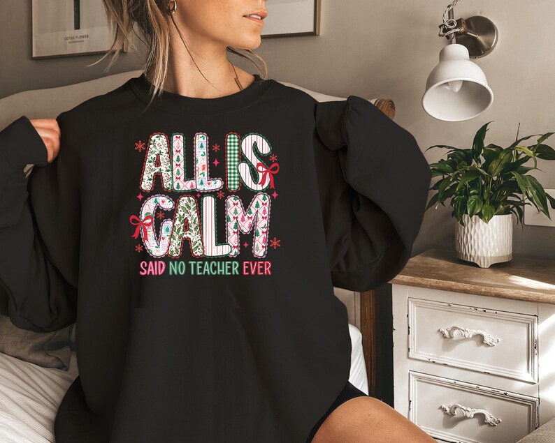 May include: Black sweatshirt with the text "ALL IS CALM" in white, green, and pink, with decorative elements. Below the text is the phrase "SAID NO TEACHER EVER" in white. The sweatshirt is a crew neck style.