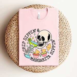 Pu&ograve; includere: Maglietta rosa con una grafica di uno scheletro che beve una margarita e il testo "Chip Dippin' & Sippin' Margarita".