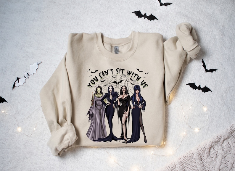 May include: A beige sweatshirt with a graphic print of four women in black dresses and the text "You can't sit with us".