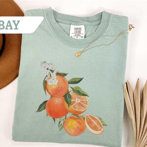 May include: A light green t-shirt featuring a watercolor illustration of oranges with green leaves and white blossoms. The shirt has a crew neck and short sleeves. A gold necklace with a small pendant is draped on the shirt. The word "BAY" is in a white banner.