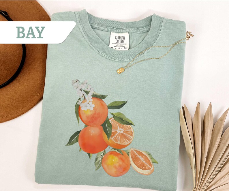 May include: A light green t-shirt featuring a watercolor illustration of oranges with green leaves and white blossoms. The shirt has a crew neck and short sleeves. A gold necklace with a small pendant is draped on the shirt. The word "BAY" is in a white banner.