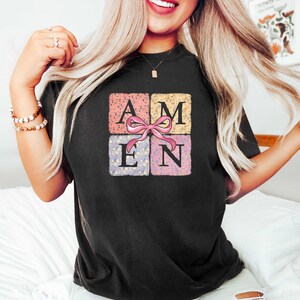 K&ouml;nnte beinhalten: Schwarzes T-Shirt mit einem floralen Design, das das Wort "AMEN" in einem Quadrat mit einer rosa Schleife zeigt.