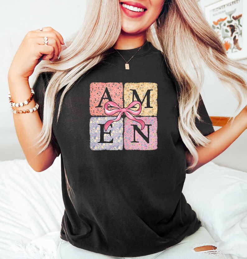 K&ouml;nnte beinhalten: Schwarzes T-Shirt mit einem floralen Design, das das Wort "AMEN" in einem Quadrat mit einer rosa Schleife zeigt.