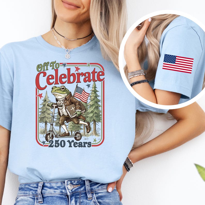 May include: Light blue t-shirt with a graphic featuring a frog riding a scooter, holding an American flag, and the text "Off To Celebrate 250 Years." An American flag patch is on the sleeve.