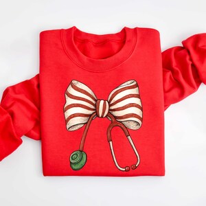 May include: A red sweatshirt featuring a whimsical design of a striped bow and a stethoscope. The bow has white and brown stripes, and the stethoscope is red with a green earpiece. The sweatshirt is a crew neck style.