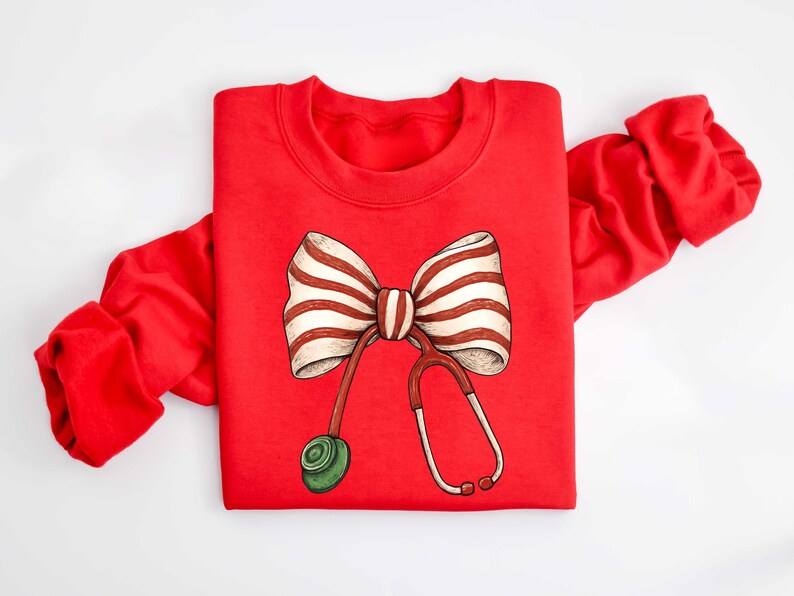 May include: A red sweatshirt featuring a whimsical design of a striped bow and a stethoscope. The bow has white and brown stripes, and the stethoscope is red with a green earpiece. The sweatshirt is a crew neck style.
