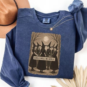 K&ouml;nnte beinhalten: Blaues Sweatshirt mit Rundhalsausschnitt und Grafik von sechs Hexen in schwarzen Kleidern und H&uuml;ten, die im Kreis stehen. Die Grafik enth&auml;lt den Text "GIRLS WILL BE GIRLS" in wei&szlig;en Buchstaben. Ein brauner Hut und eine goldene Halskette sind ebenfalls sichtbar.