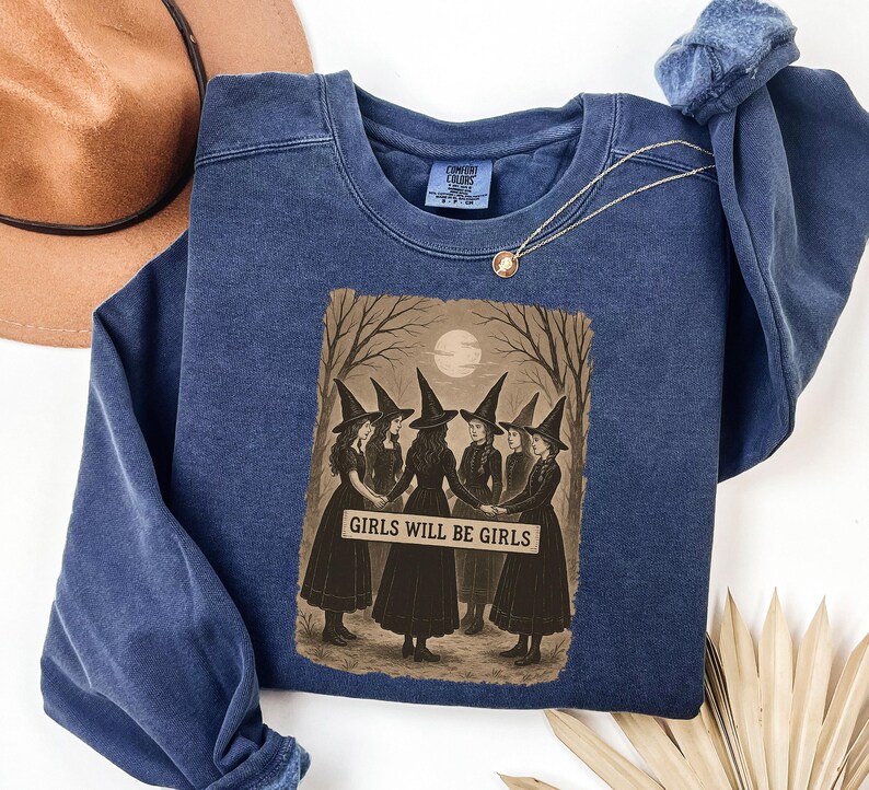 K&ouml;nnte beinhalten: Blaues Sweatshirt mit Rundhalsausschnitt und Grafik von sechs Hexen in schwarzen Kleidern und H&uuml;ten, die im Kreis stehen. Die Grafik enth&auml;lt den Text "GIRLS WILL BE GIRLS" in wei&szlig;en Buchstaben. Ein brauner Hut und eine goldene Halskette sind ebenfalls sichtbar.