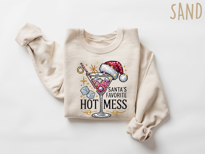 May include: A beige sweatshirt with a festive design featuring a martini glass with a red liquid, ice cubes, and a red Santa hat. The text "Santa's Favorite Hot Mess" is printed in gold glitter.