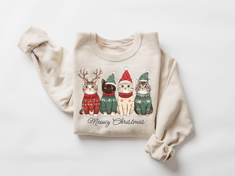 May include: A beige sweatshirt with a Christmas-themed design featuring four cats wearing Santa hats and sweaters. The text "Meowy Christmas" is printed below the cats.
