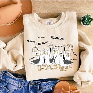 May include: Cream-colored sweatshirt featuring a cartoon illustration of seagulls with the text "MS. JACKIE!". The seagulls are in various poses, with black and white details. The sweatshirt is paired with blue denim shorts and sandals.