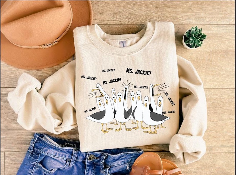 May include: Cream-colored sweatshirt featuring a cartoon illustration of seagulls with the text "MS. JACKIE!". The seagulls are in various poses, with black and white details. The sweatshirt is paired with blue denim shorts and sandals.