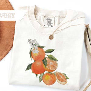 May include: Ivory-colored t-shirt with a watercolor design of oranges, green leaves, and white flowers. The shirt has a crew neck and short sleeves. A gold necklace with a pendant is draped on the shirt.
