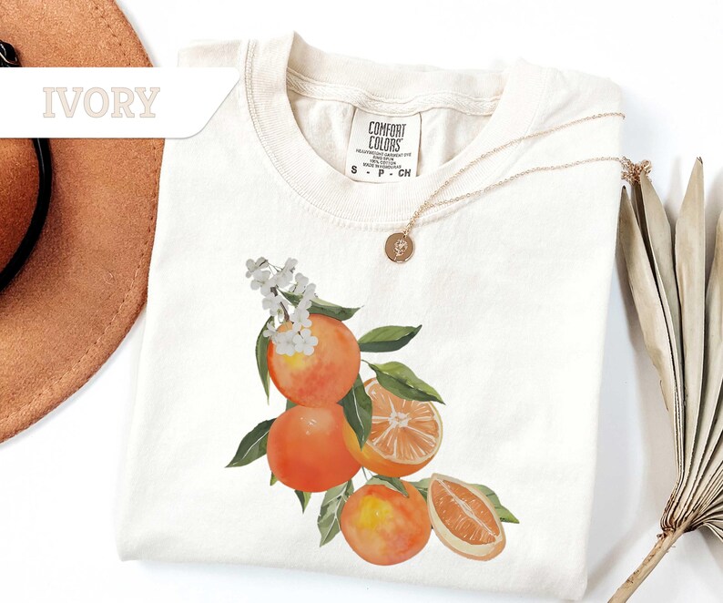 May include: Ivory-colored t-shirt with a watercolor design of oranges, green leaves, and white flowers. The shirt has a crew neck and short sleeves. A gold necklace with a pendant is draped on the shirt.