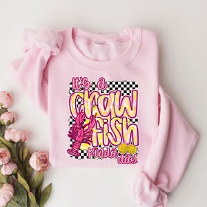 Puede incluir: Sudadera rosa con un dise&ntilde;o gr&aacute;fico de un cangrejo de r&iacute;o y el texto "It's a crawfish kinda day".